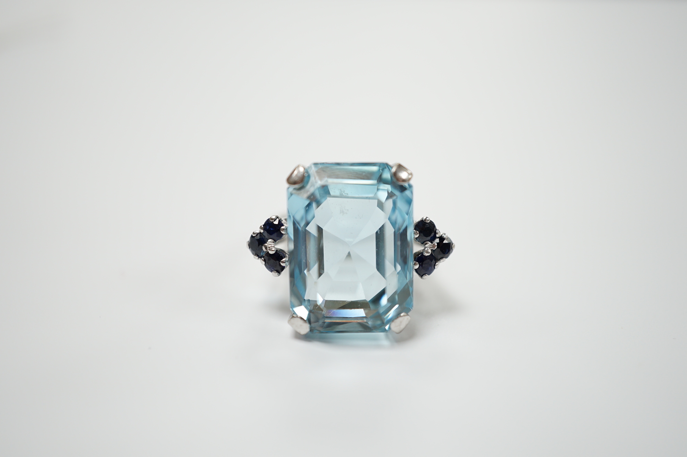 An 18ct white metal and single stone emerald cut aquamarine set dress ring, with six stone sapphire set shoulders, size L, gross weight 11.8 grams.